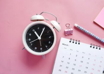 Pembe arka plan üzerinde çalar saat, takvim yaprağı ve tükenmez kalem yer alıyor.