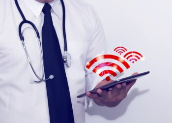 Doktor önlüklü bir kişi elinde tablet tutuyor ve üzerinde kablosuz ağ sembolleri var.