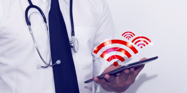 Doktor önlüklü bir kişi elinde tablet tutuyor ve üzerinde kablosuz ağ sembolleri var.
