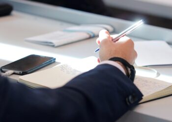 Masada oturan bir kişi deftere not alıyor ve akıllı telefon kullanıyor.