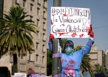 Sağlık çalışanı önlüklü bir protestocu hastane önünde pankart tutuyor.
