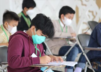 Sınıfta maske takarak sınavda oturan öğrenciler ve başlarında duran gözetmen öğretmen.