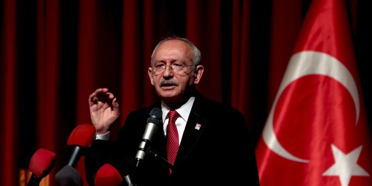 Kırmızı fon önünde mikrofonla konuşan bir siyasetçi ve Türk bayrağı.