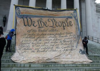Tarihi bir binanın merdivenlerinde We The People yazılı büyük anayasa metni afişi.