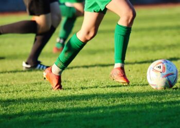 Çim sahada top süren ve mücadele eden futbolcuların bacakları ve kramponları.