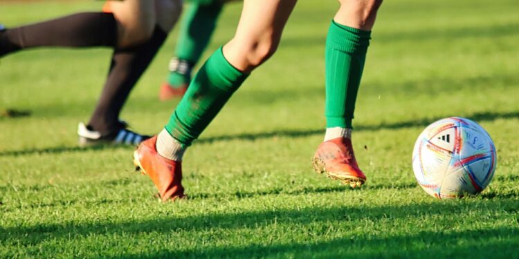 Çim sahada top süren ve mücadele eden futbolcuların bacakları ve kramponları.