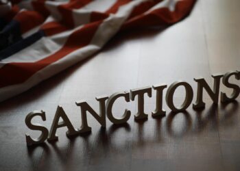 Ahşap bir zeminde İngilizce 'Sanctions' yazısı ve arkasında Amerikan bayrağı duruyor.