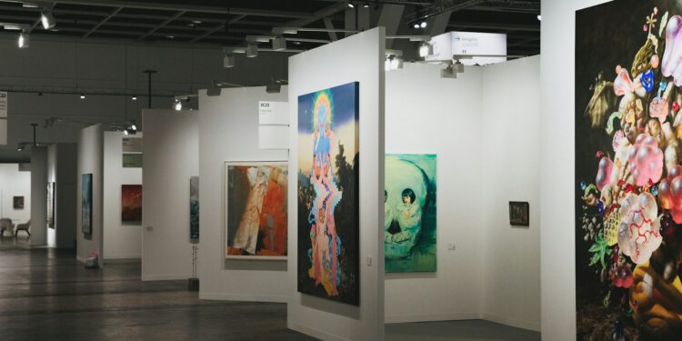 Geniş beyaz duvarlı bir çağdaş sanat fuarında sergilenen büyük tablolar.