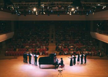 Devasa konser salonunda sahnede piyano eşliğinde çalan müzisyenler ve izleyiciler.