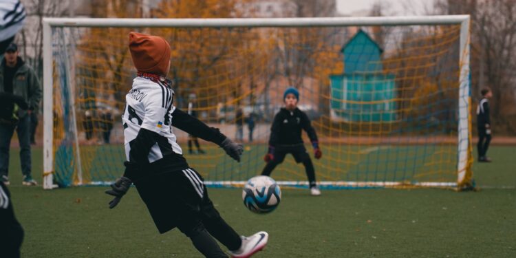 Futbol sahasında fileler önünde topa vuran sporcu ve kaleci